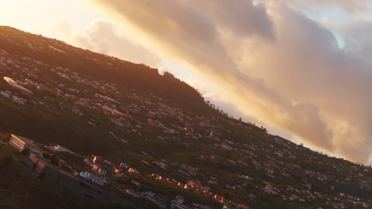 Tilted drone shot flying over Calheta village on hillside during golden hour sunset, warm light illuminates clouds and landscape on Madeira island, Portugal. Aerial drone