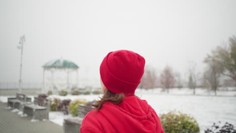 lady jogging along snowy pathway wearing red beanie and jacket, hair fluttering in cold breeze, surrounded by benches, colorful bushes, gazebo, and serene winter scenery with soft foggy atmosphere