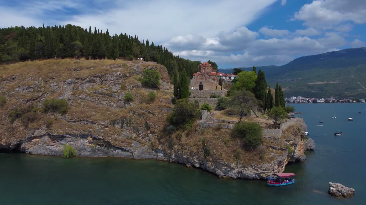 Cinematic shot of iconic Orthodox Church on Ohrid peninsula with boat sailing along turquoise waters - Macedonia