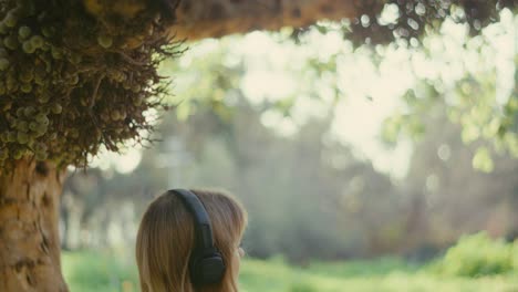 lady with loose hair sits on bench under tree listening to music on headphones