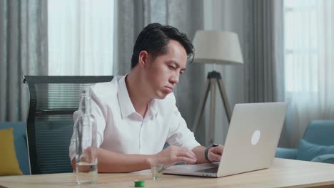 아시아인 남자가 술을 마시기 전에 글라스에 보드카를 부어 집에 있는 노트북에 타이핑하고 있다