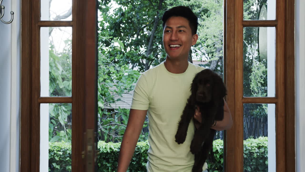 Asian man entering home holding dog, smiling warmly at doorway