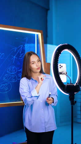 lady blogger taking video on her phone on tripod with light ring. woman talks, points at numbers on the glass wall and gestures. vertical video.