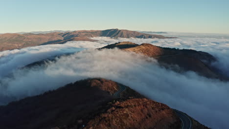 Majestic-mountain-peaks-piercing-through-a-sea-of-clouds-at-sunrise,-winding-road-visible