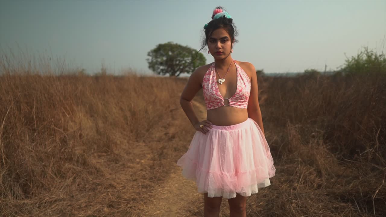 una atractiva mujer asiática con un vestido de tutú rosa, empujando su cabello hacia atrás sobre sus orejas mientras está de pie en un campo de hierba seca rodeado de naturaleza, india