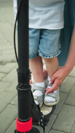 close-up of a woman's hand guiding a young boy's foot onto the scooter platform as he lifts his left leg