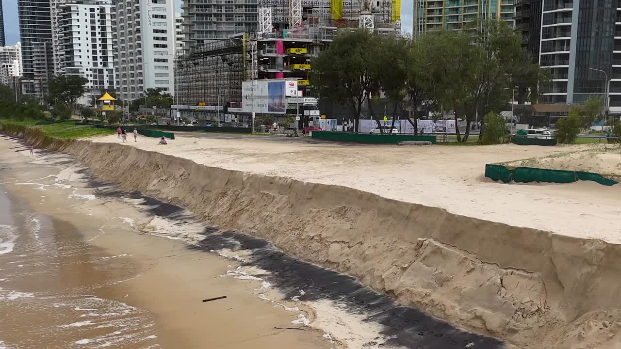 Toma de dron mostrando olas del océano rompiendo contra acantilados de arena erosionados frente a rascacielos de la ciudad.