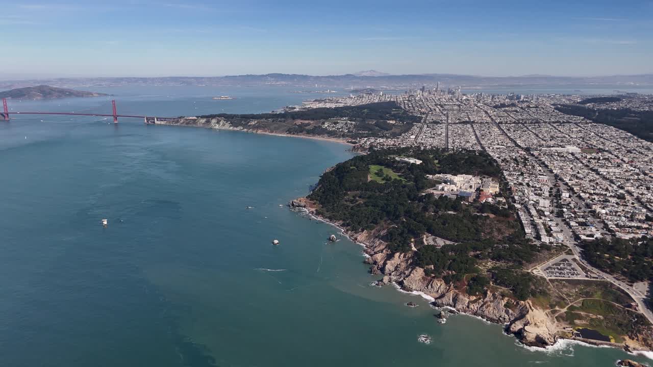 Drone photo of San Franciscoâ€™s rugged coastline with the Golden Gate Bridge, lush greenery, and urban layout under a blue sky. Alcatraz visible in the bay.