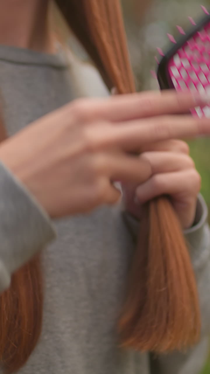 Close-up of woman gently brushing her long, straight reddish-brown hair with a pink hairbrush, focusing on the smooth texture and care in a serene forest setting