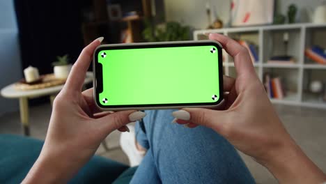 woman using smartphone with green screen. unrecognizable female person holding mobile phone with chroma key, sitting on sofa in living room. watching film or video. spare time at home
