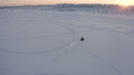 drone shot of a fast snowmobile rides towards the sunset and forest during a cold winter season in sweden