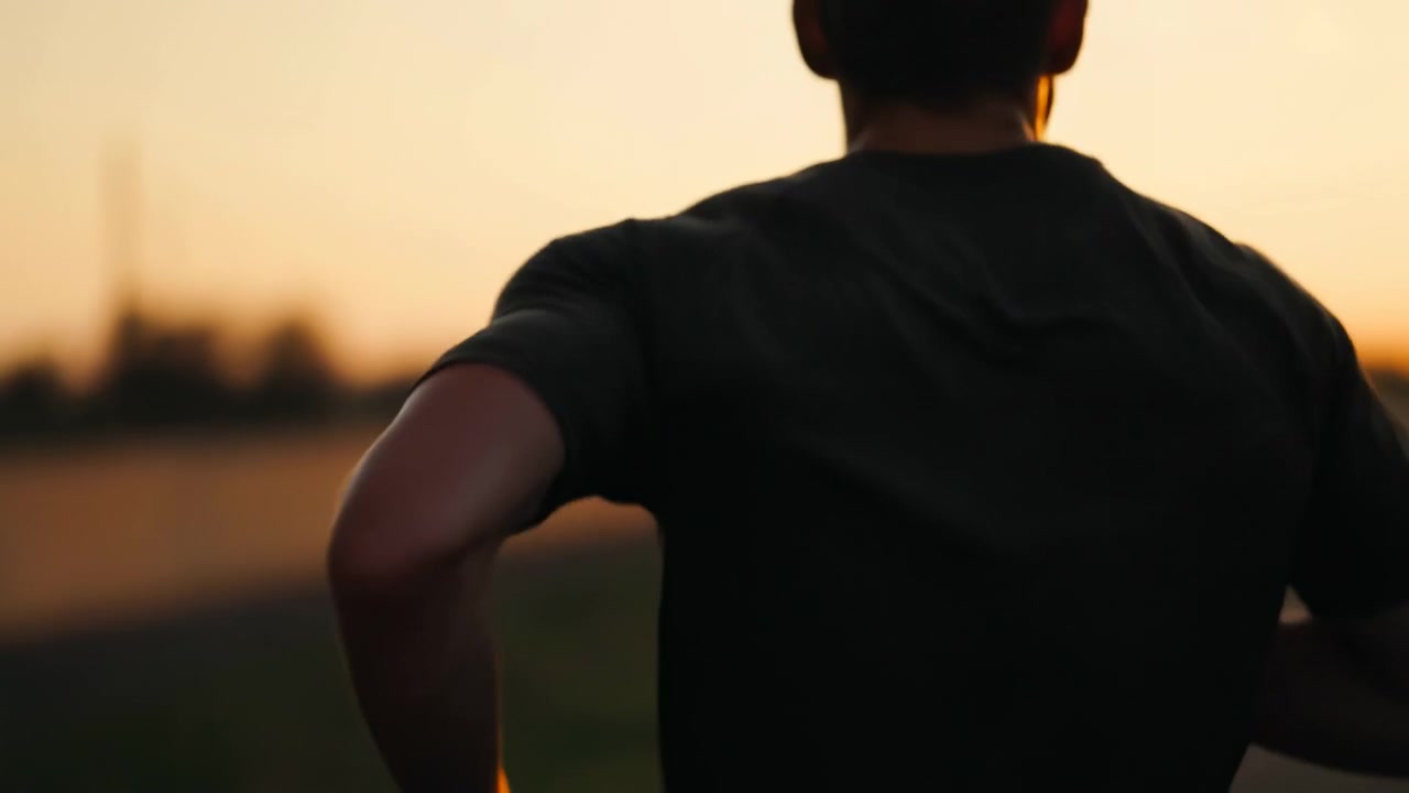 Rear view of male runner moving along roadside during golden hour, with bent arms, forward lean, and torso rotation creating sense of ongoing outdoor fitness activity against warm sky