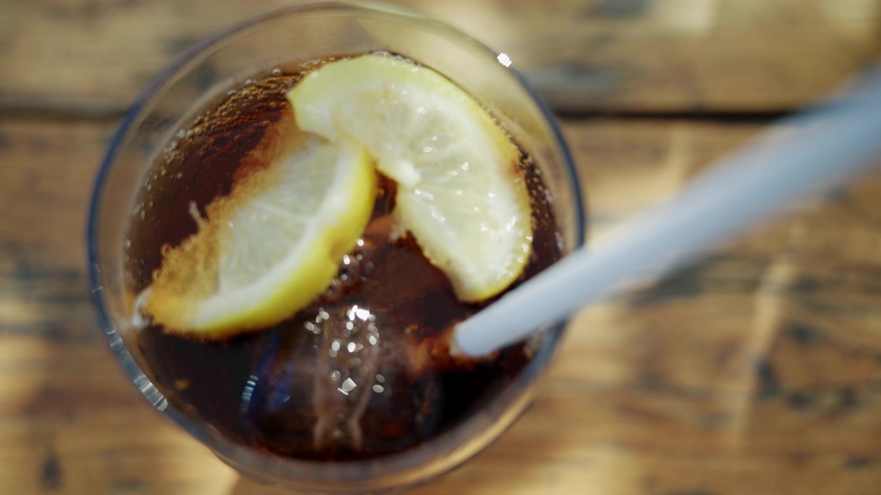 Primo piano di una bevanda cola ghiacciata con fette di limone mescolata con una cannuccia bianca in un bicchiere trasparente.