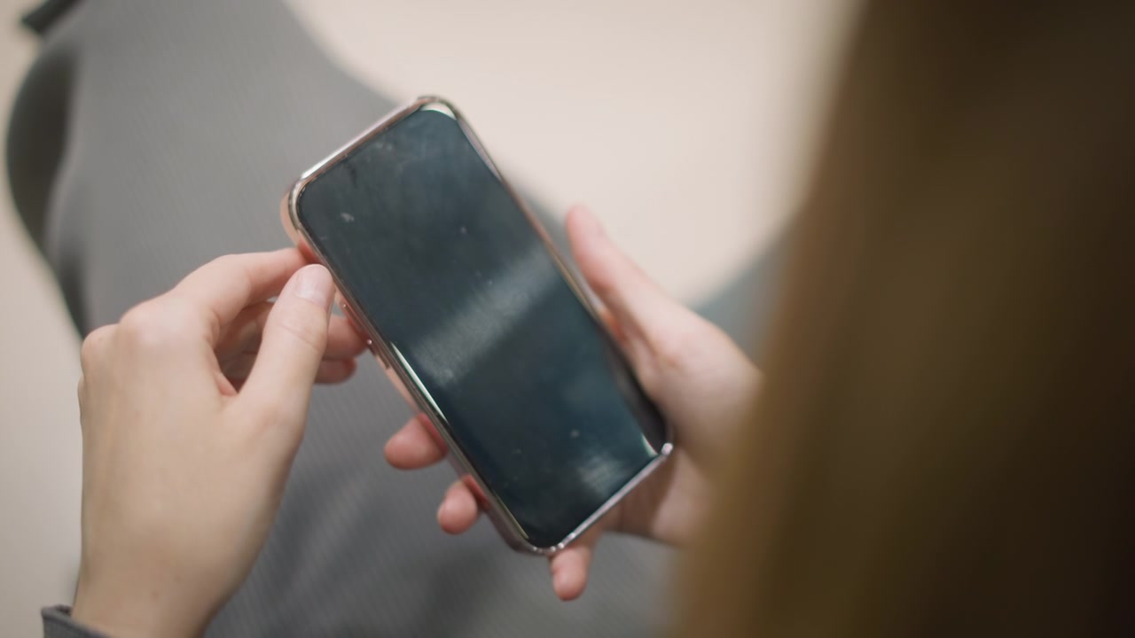 Primo piano di una donna che tiene uno smartphone tra le mani. Il dispositivo è tenuto saldamente, mostrando il suo schermo che riflette la luce, mentre la donna lo manipola delicatamente. Un'illuminazione soffusa crea.