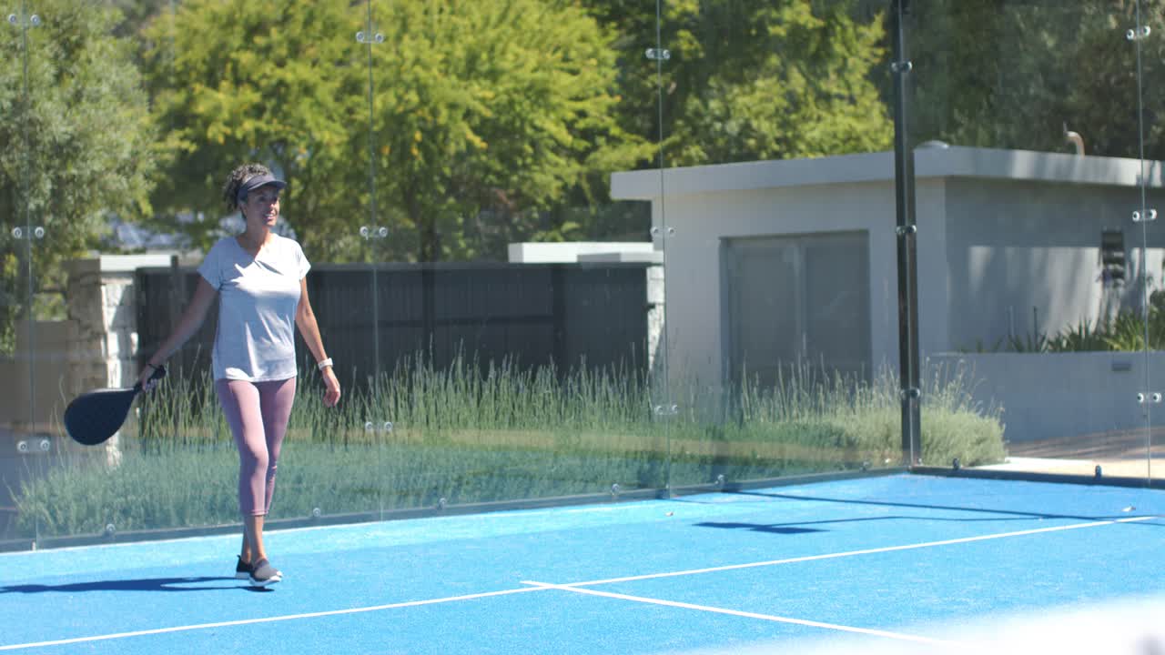 African American woman tossing ball, serving with paddle on blue court, wearing visor for practice