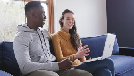 una pareja feliz y diversa sentada en el sofá, usando una computadora portátil y una tarjeta de crédito en casa, espacio para copiar
