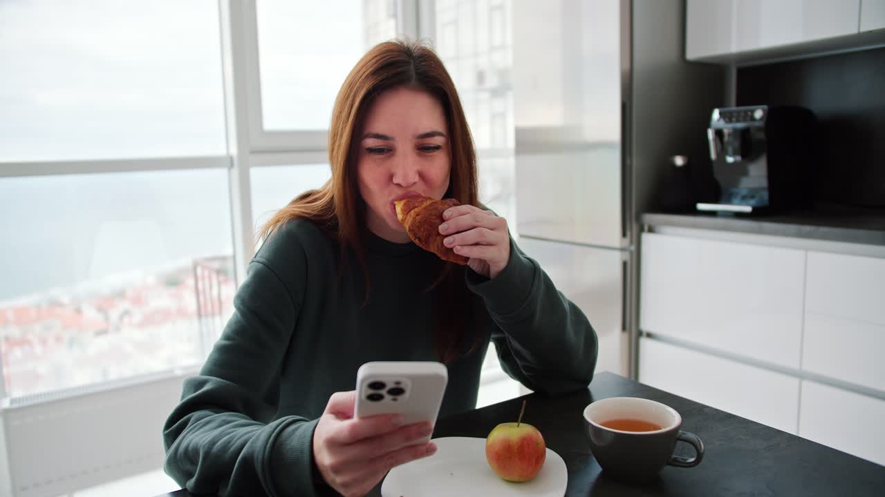 feliz y segura chica morena con un suéter verde come un croissant y té durante su desayuno y navega por las redes sociales en su teléfono inteligente blanco en un apartamento moderno en la cocina
