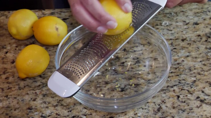 Una donna che grattugia la scorza di limone con una grattugia di metallo in una ciotola di vetro trasparente