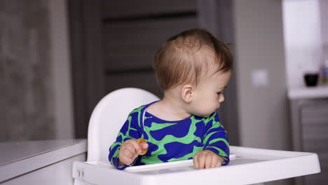cute toddler boy in blue and green shirt sitting at feeding table. mother is giving a full spoon to her child and he takes it. blurred backdrop.