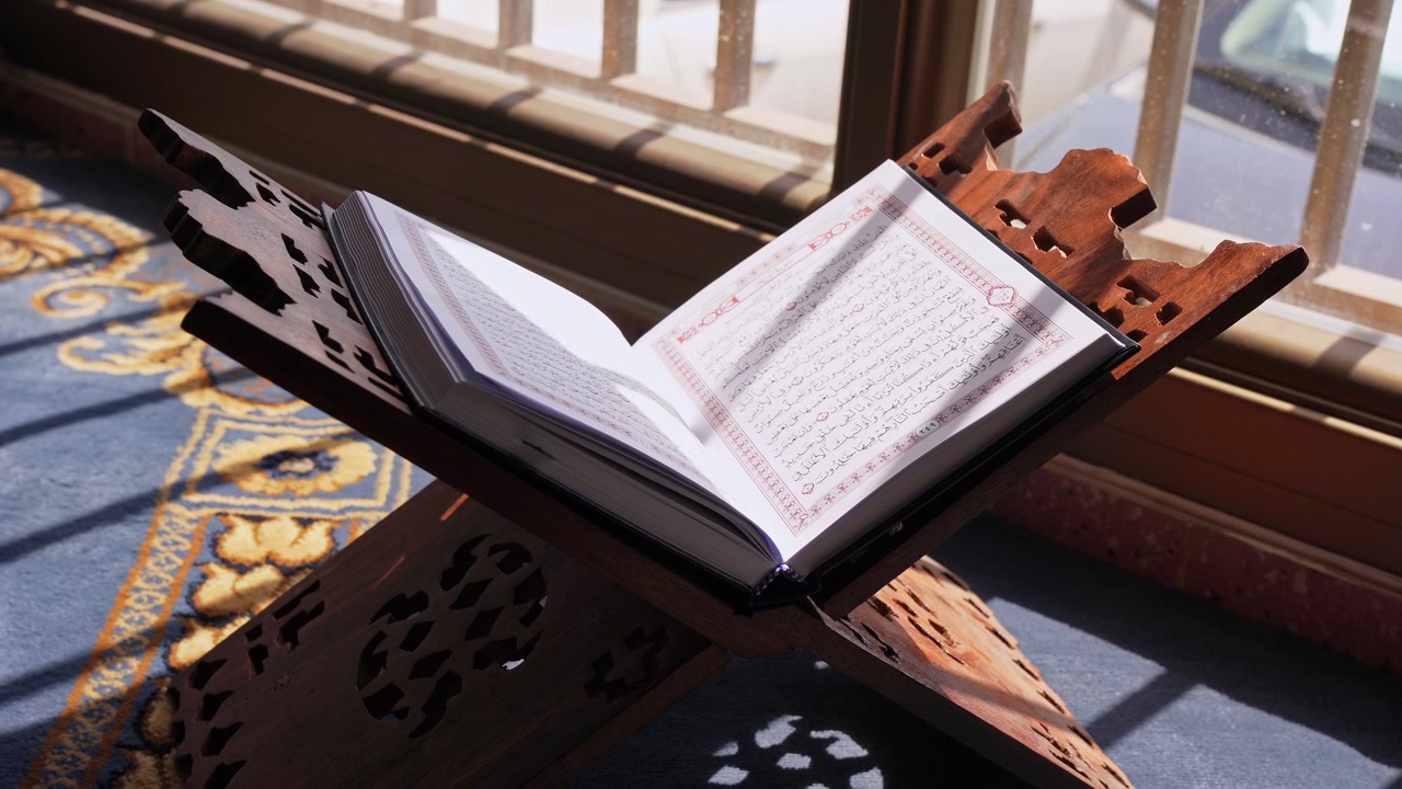 Un Sacro Corano aperto poggia su un leggio in legno intagliato accanto a una finestra luminosa. La forte luce del giorno proietta ombre drammatiche sul testo arabo e sul tappeto blu a motivi della moschea durante una mattinata tranquilla.