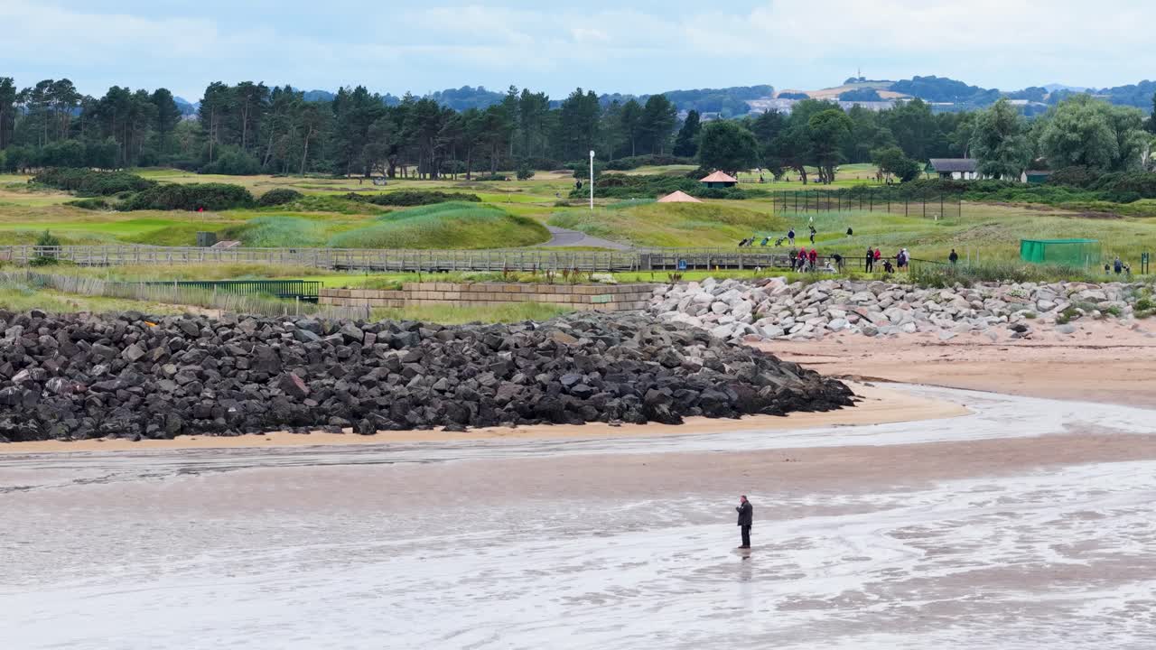 Single person walks along wet sand, golf course and rock wall in background, overcast daylight