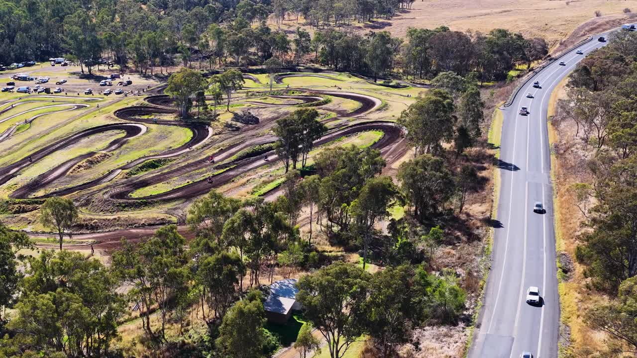 Drone footage shows vehicles traveling on rural road beside motocross track, bright daylight, steady shot