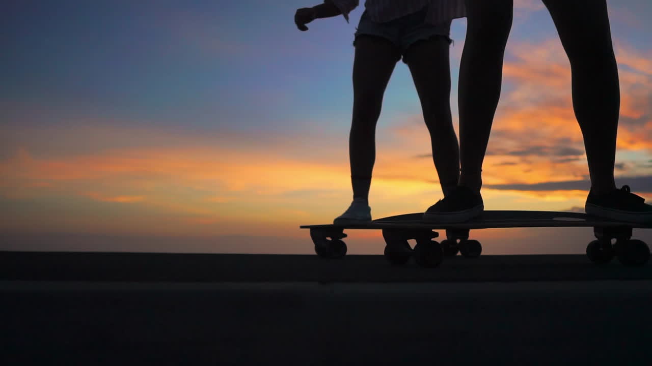 Skateboarding on a road at sunset, two friends create a mesmerizing scene in slow motion with mountains and a beautiful sky. They're attired in shorts