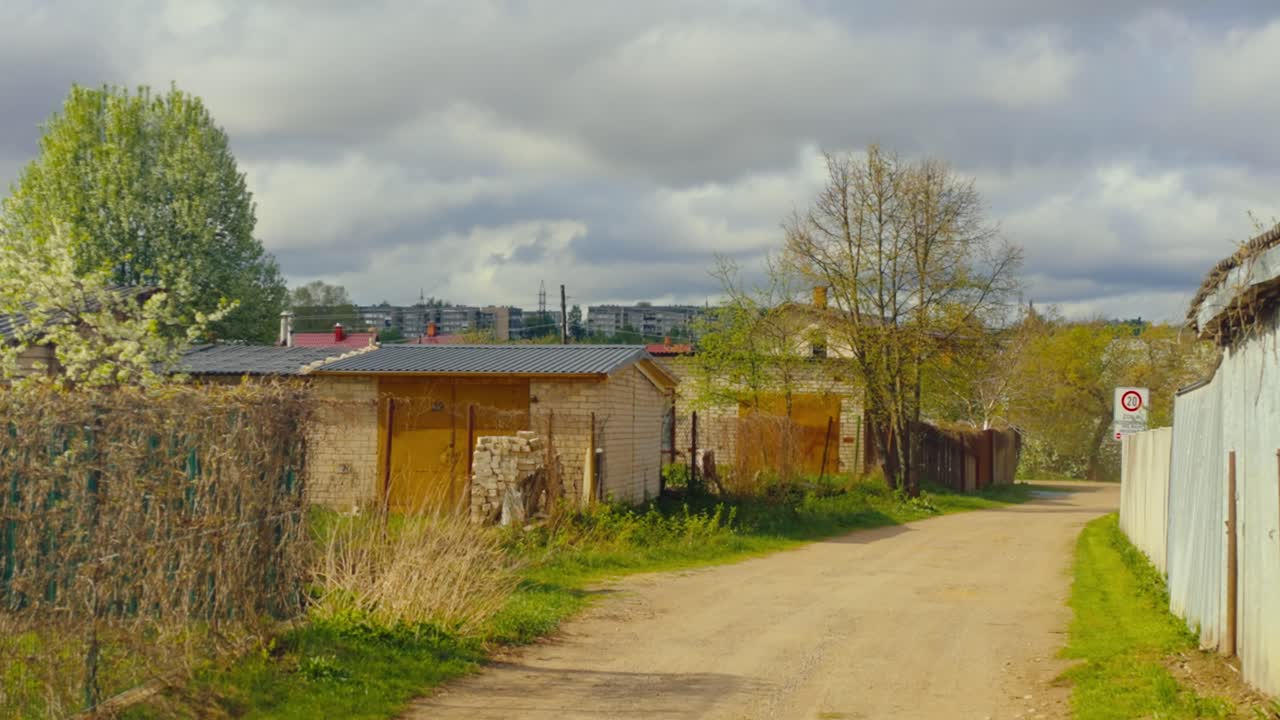 A rural dirt road lined with small garages, fences, dry grass, and spring trees under a bright cloudy sky. Daugavpils, Latvia (Latgale)