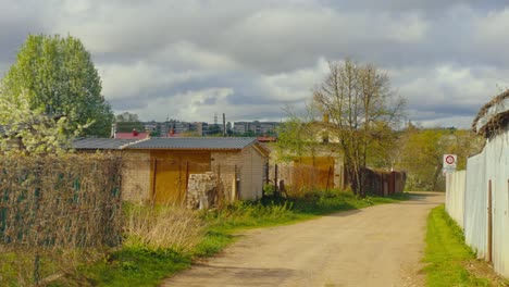 a rural dirt road lined with small garages, fences, dry grass, and spring trees under a bright cloudy sky. daugavpils, latvia (latgale)