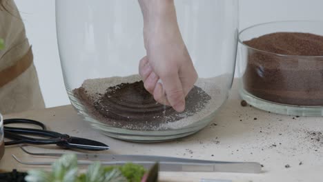 the woman's hand puts the sand into the huge glass terrarium to create a live tiny environment concept close-up