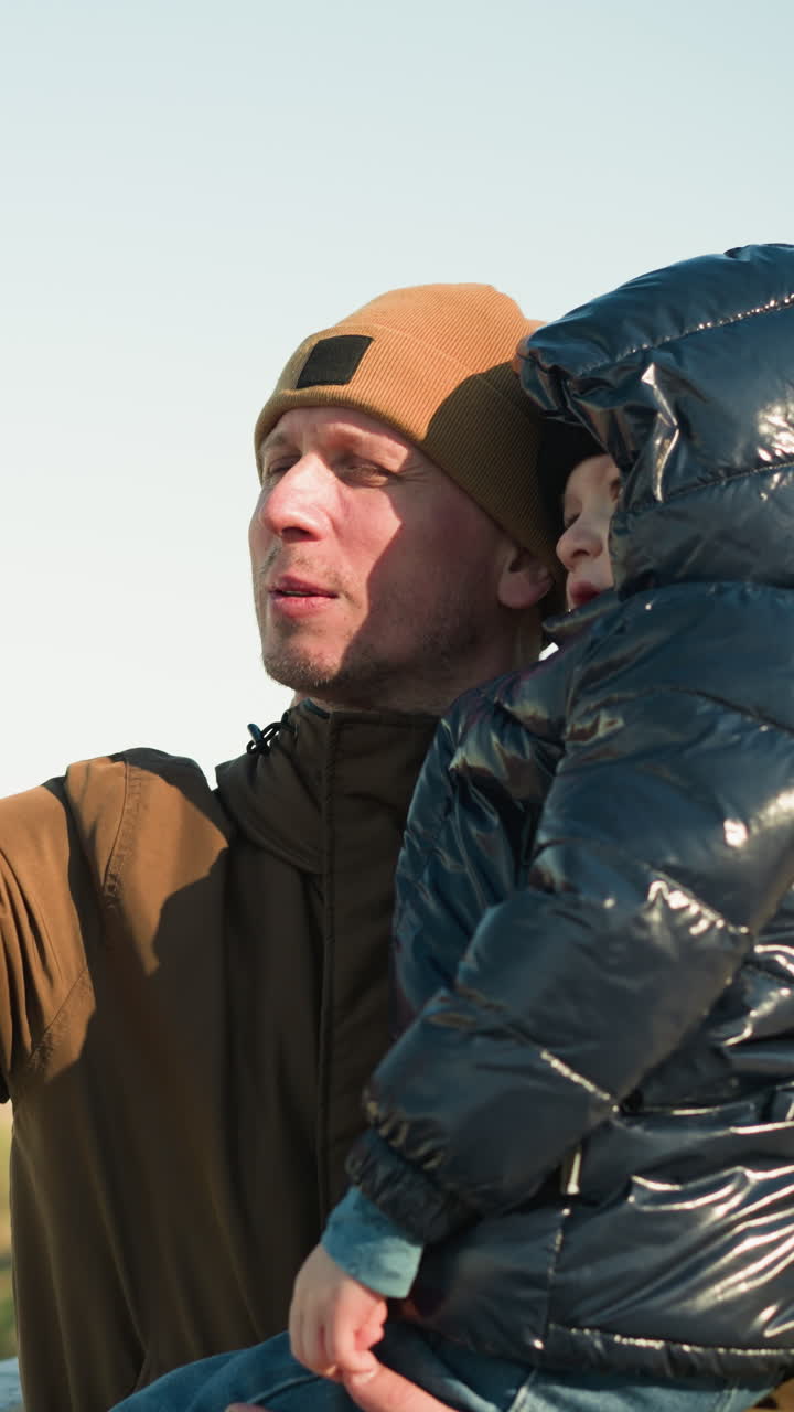 한 아버지가 아름다운 전망대 근처에 서있는 동안 아들을 들고 있습니다. 갈색 재과 모자를 입은 아버지는 검은색 재을 입은 아이에게 무언가를 설명하고 있습니다.