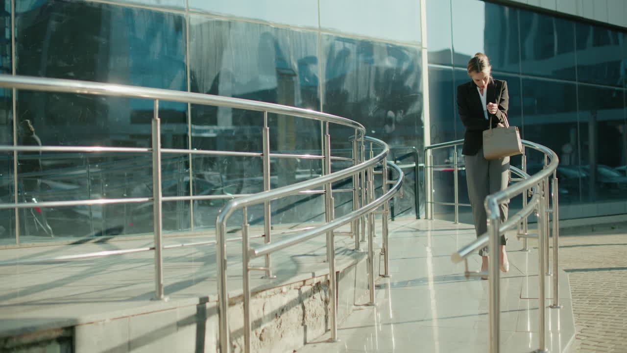 Professional businesswoman walking down ramp near modern glass building, thoughtfully checking handbag, reflections of environment visible