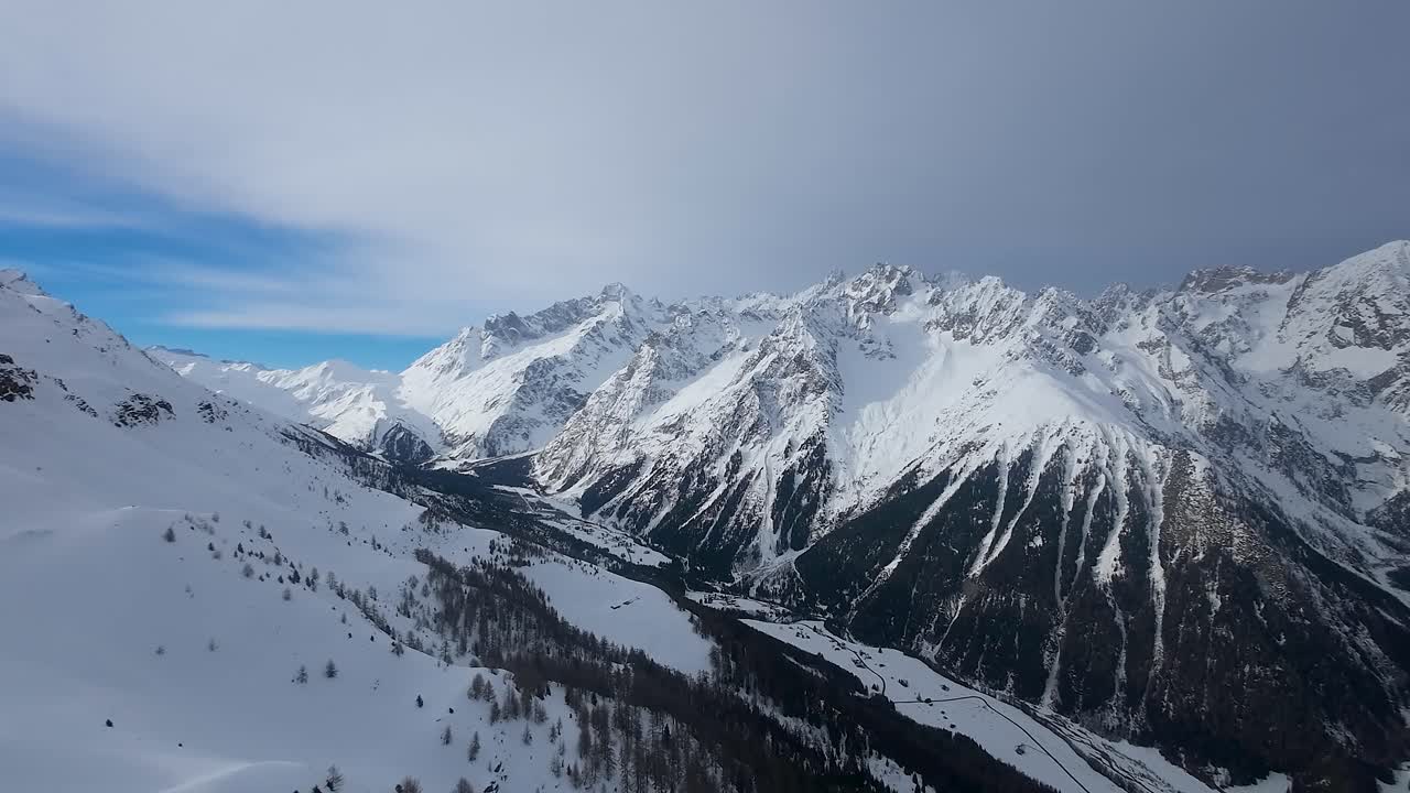Ripresa drone con effetto parallasse di una catena montuosa innevata con cime ripide e aspre sotto un cielo nuvoloso.