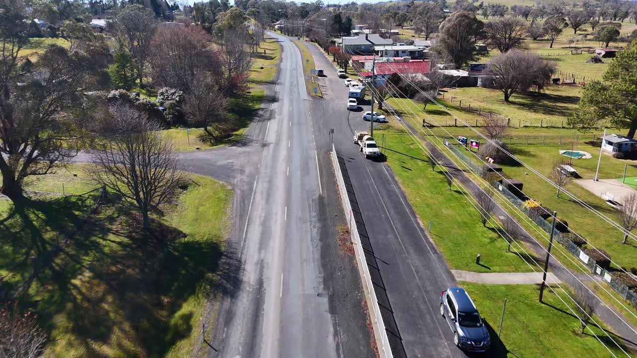 Drone footage follows a quiet residential street in a rural Australian town, showing houses, parked cars, and green lawns under bright natural sunlight