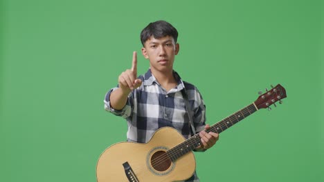 녹색 스크린 배경에 서 있는 동안 기타를 가진 아시아 십대는 제스처를 표시하지 않습니다.