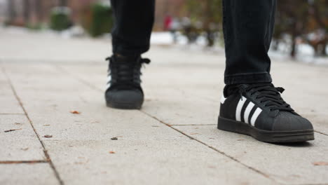 close-up leg view of individual in black trousers and sneakers with white stripes walking on an interlocked path, blurred background with snow, trees, and an unclear figure