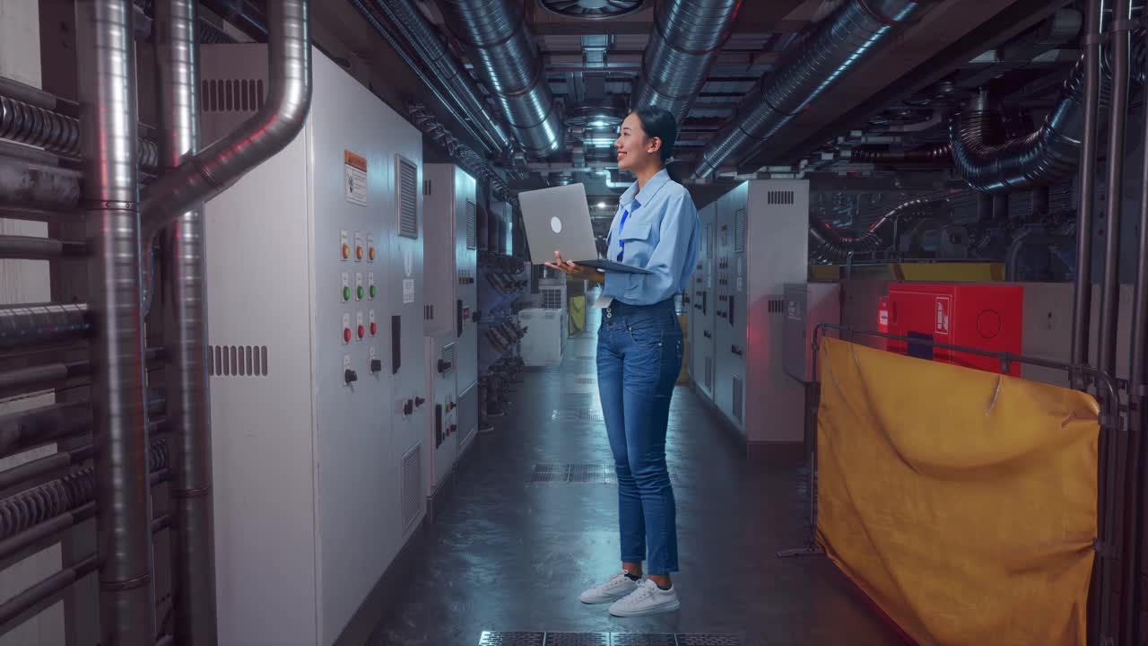 Full Body Side View Of An Asian Female Worker Standing In Engine Control Room, Observes By Looking Up Before She Come To Concentrating On The Laptop And Keep On Typing