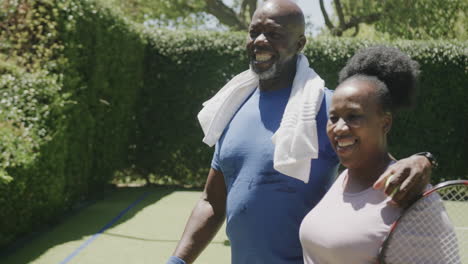 happy senior african american couple walking with tennis rackets and embracing at tennis court