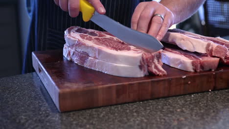 caucasian male hands slice and cut raw meat with large sharp yellow knife on wood chopping block in kitchen to make sausage, static close up