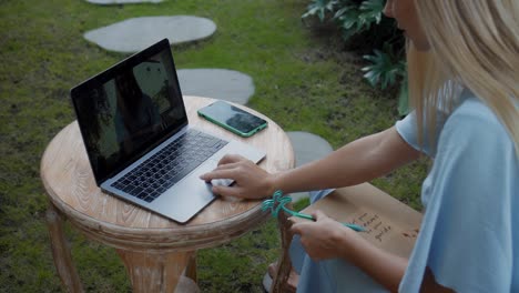 una chica en un vestido azul remotamente en línea trabajando detrás de una computadora portátil y mirando a la pantalla el patio trasero con plantas verdes