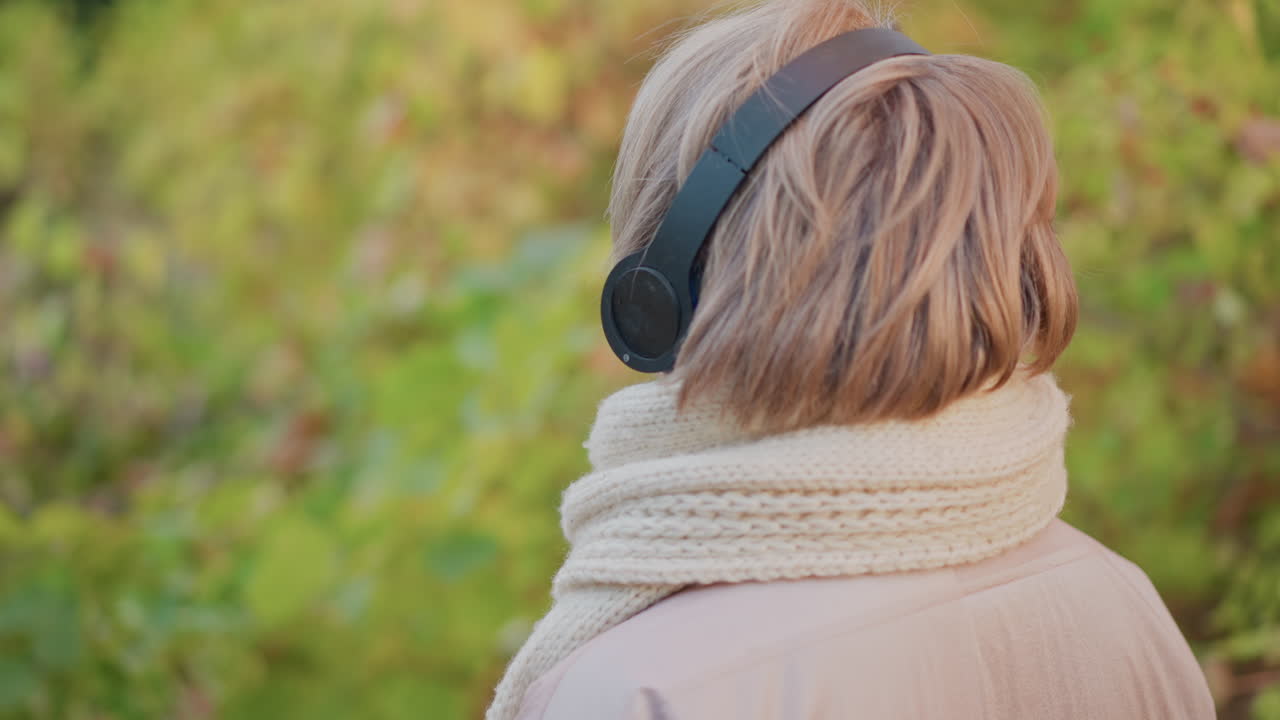 rear view woman wearing headphones walking through autumn forest path, knitted scarf and padded jacket visible, golden leaves carpeting ground, soft light filtering through trees, peaceful solitary stroll