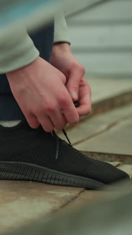 primer plano de alguien atando su cordón de zapatos en una escalera oxidada, con una vista borrosa de alguien con un suéter marrón caminando en el fondo