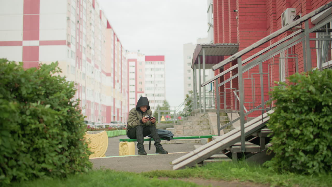 Young kid seated alone on outdoor bench wearing hooded jacket focused on gaming device with backpack placed beside, surrounded by greenery, leaves swaying gently, parked cars and tall apartment