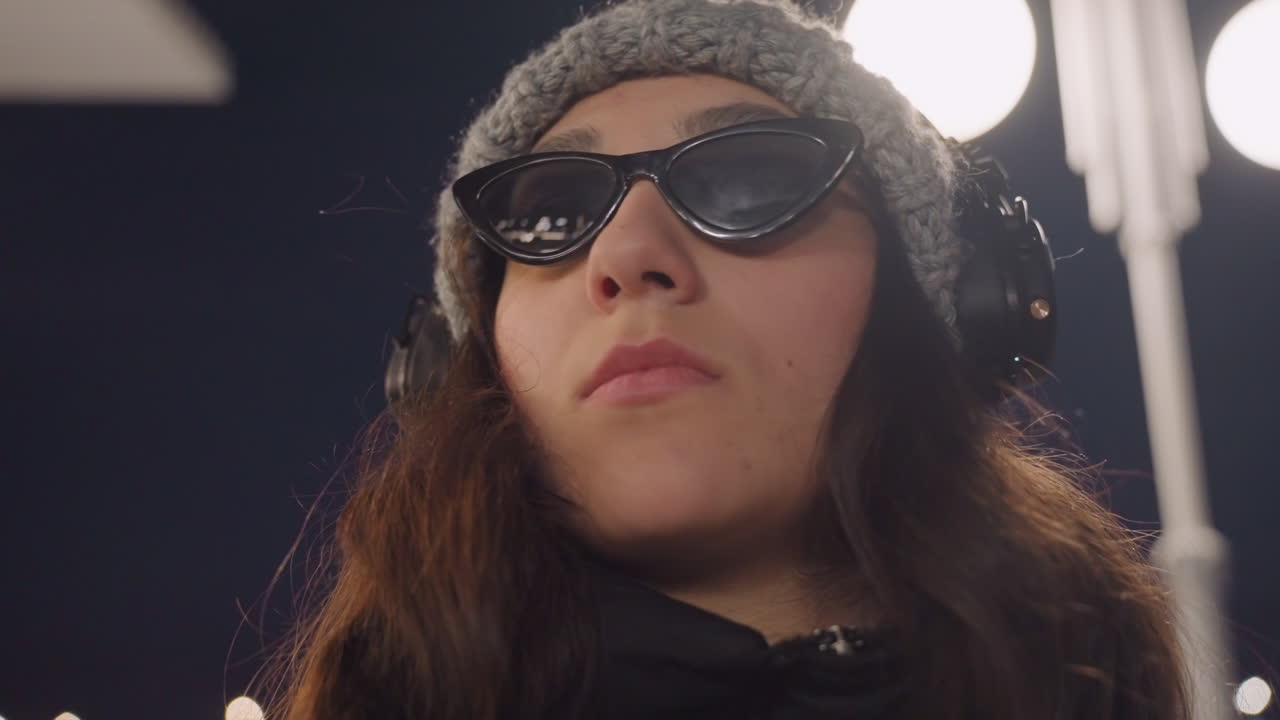 Nattens gade, danseportræt med hovedtelefoner, der viser ung kvinde iført solbriller og hue, lavvinklet filmisk nærbillede, gadelysets skær, selvsikkert udtryk med diskret hovedbevægelse, lofi bystemning.