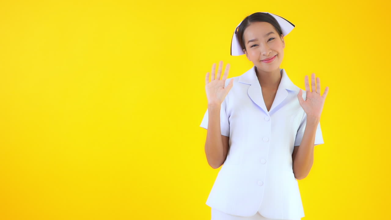 Static slow-motion shot of a cute Asian nurse, waving both hands and smiling cheerfully in front of a yellow background