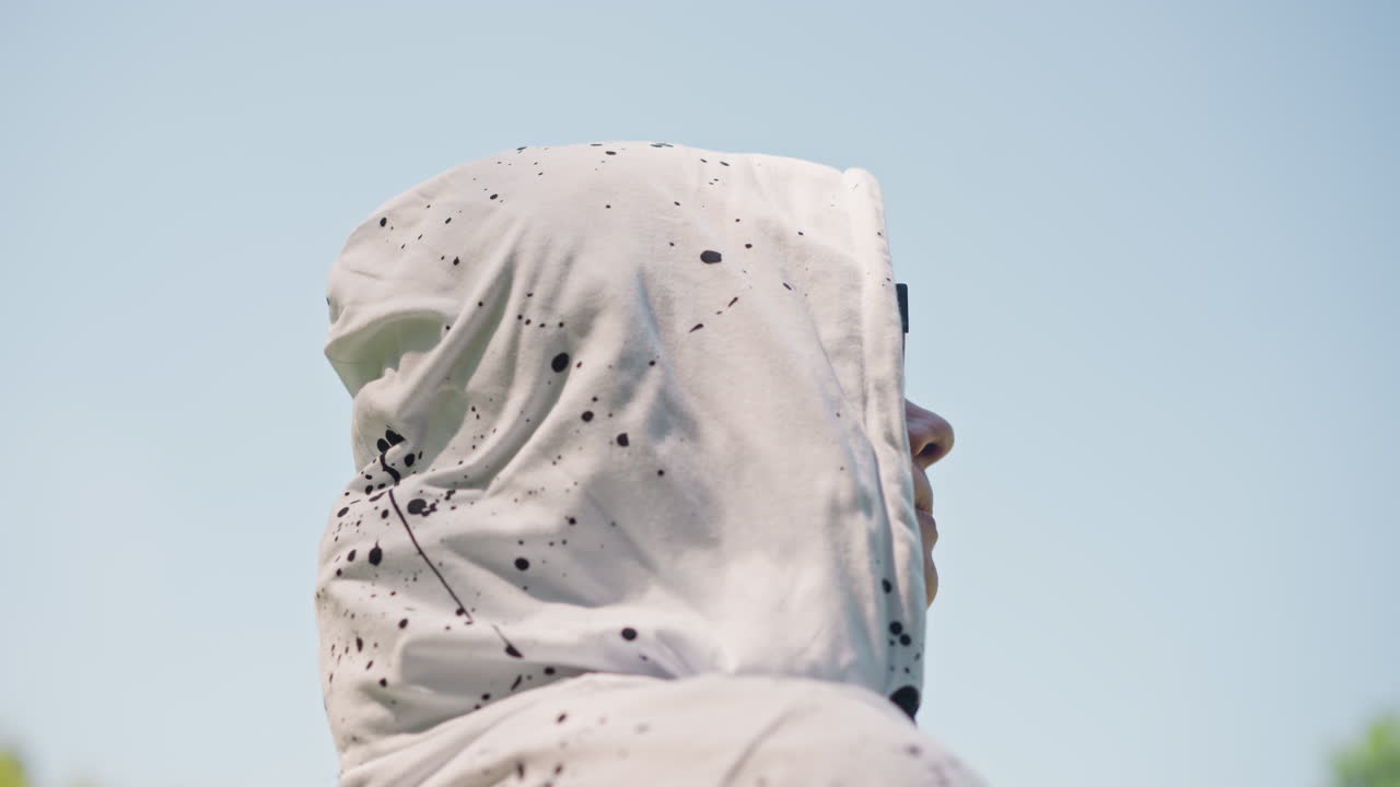 Sort kvinde med hvid hættetrøje, profil udendørs, eftertænksom. Studerende justerer hætten, mens hun står i en solbeskinnet park, brillerne reflekterer den blå himmel, malingsstænk på stoffet, roligt og tankefuldt udtryk.