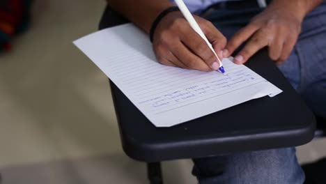 a top close up view of a boy student writing on the paper at his class room, wearing a jeans pant, a chair with its writing desk