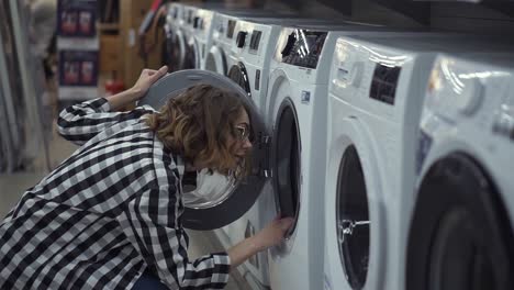 a young positive woman in a plaid shirt choosing washing machine in the shop of household appliances. opens the door and look inside. side view. slow motion