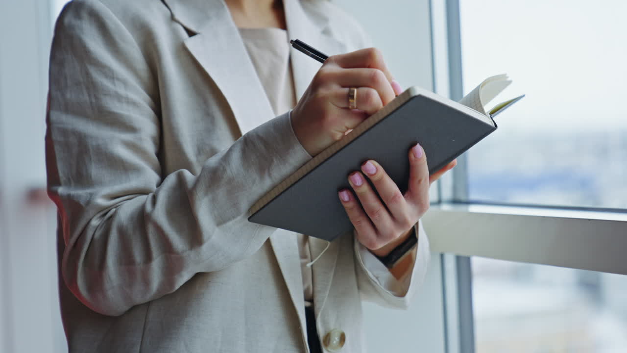 Woman in light jacket standing at the big window. Lady holding a notebook, writing something down there and closing it. Close up.