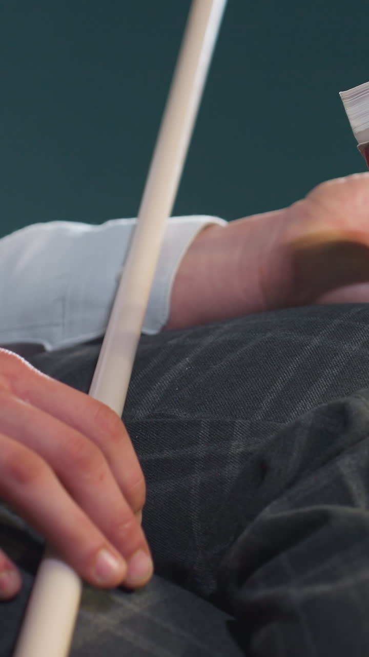 색 셔츠와 회색 바지를 입은 얼굴이 없는 사람이 책과 큐 스틱을 들고 있는 클로즈업. 손은 무에 앉아 손가락으로 큐스틱을 잡고 있다. 부드러운 조명은 옷과 물건의 질감을 강조한다.